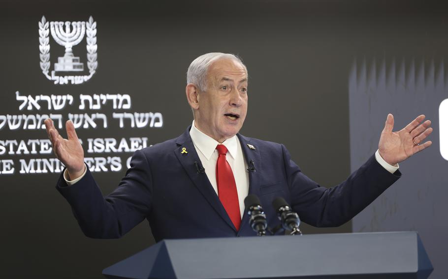 Benjamin Netanyahu speaks at a podium while raising both of his hands.