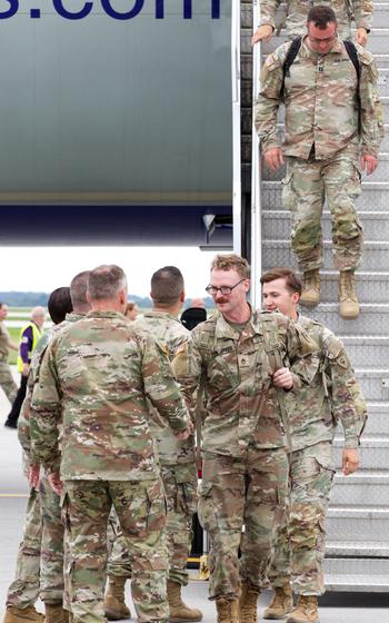 A vertical photo of soldiers walking down the steps of an airplane, with one of the soldiers shaking hands with others waiting at the bottom of the steps.