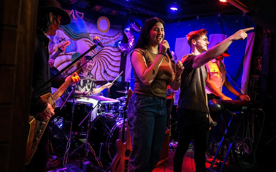A woman sings into a microphone while a man stands next to her and points outward while standing on a stage with a drummer behind them and a guitarist to the side.