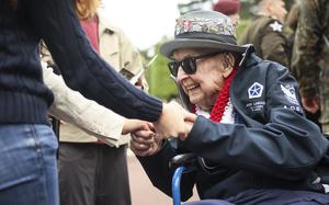 An elderly military veteran in a wheelchair wearing sunglasses and a military cap adorned with multiple service pins shakes hands with someone in a navy blue suit jacket. The veteran is wearing a navy blue jacket with military patches and a red, white and blue lei around his neck. Other veterans and attendees can be seen in the blurred background.