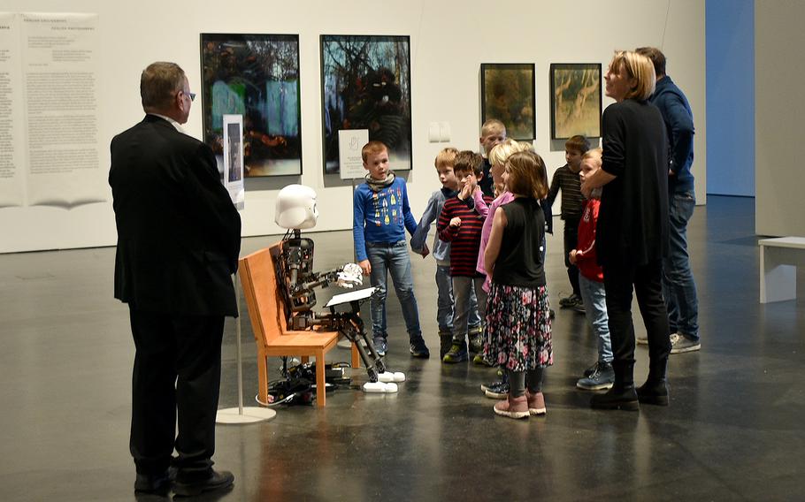 A group gathers around Robot Yeong-Hee, a robot installation that is part of the “Kim Soun-Gui: Lazy Clouds” exhibit, on Nov. 26, 2022, at the Zentrum fuer Kunst und Medien Karlsruhe in Karlsruhe, Germany. Missing from the installation is a tree, which was “getting fresh air.”