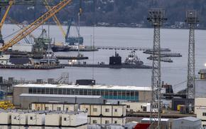 An aerial view of a shipyard along the banks of a body of water, with the top of a submarine visible above the water line surrounded by cranes.