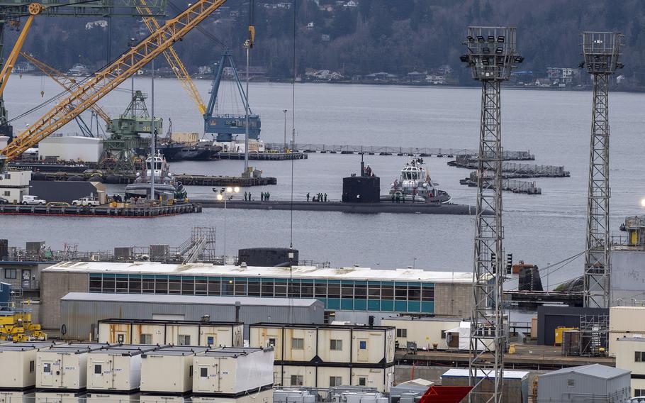 An aerial view of a shipyard along the banks of a body of water, with the top of a submarine visible above the water line surrounded by cranes.