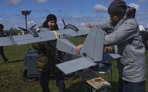 Two men examine a gray military-style drone at an outdoor event. They are handling the unmanned aircraft, which has multiple propellers. The setting is a grassy field under a partly cloudy blue sky, with other people and equipment visible in the background.
