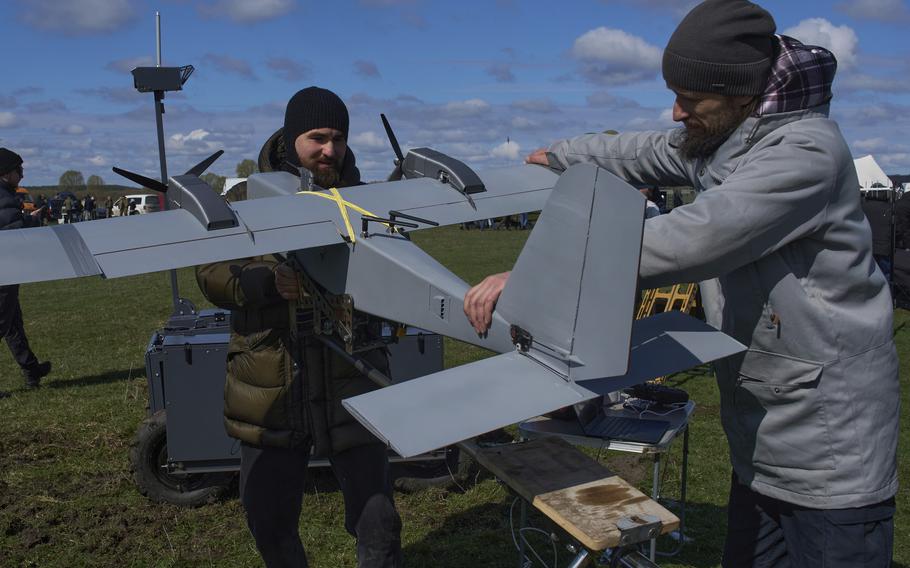Two men examine a gray military-style drone at an outdoor event. They are handling the unmanned aircraft, which has multiple propellers. The setting is a grassy field under a partly cloudy blue sky, with other people and equipment visible in the background.