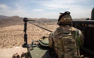 An Army soldier in camouflage uniform and helmet, seen from behind looks out over an expanse of desert land, with mountains and the horizon in the background.