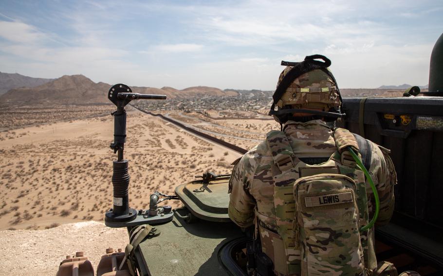 An Army soldier in camouflage uniform and helmet, seen from behind looks out over an expanse of desert land, with mountains and the horizon in the background.