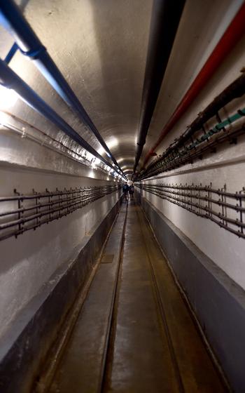 Long underground tunnel with railway tracks running down the center, industrial piping along the ceiling, and lighting extending into the distance.