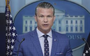Pete Hegseth, wearing a blue suit and seen from the chest up, speaks into two microphones while standing in front of a U.S. flag and the emblem of the White House behind him.