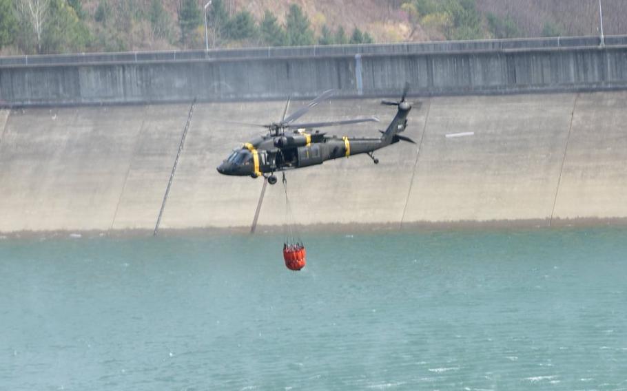 A military helicopter hovers in the air as it lowers a red bucket underneath it to collect water from a reservoir.