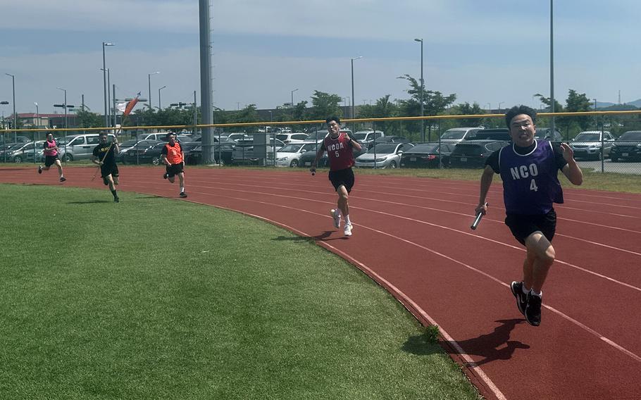 U.S. and South Korean soldiers in exercise clothing run a relay race on a track with a fence in the background and parked cars in a lot beyond it.