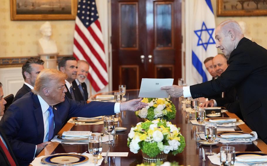 Benjamin Netanyahu hands Donald Trump a folder across a large conference table with other officials seated behind them at place settings.