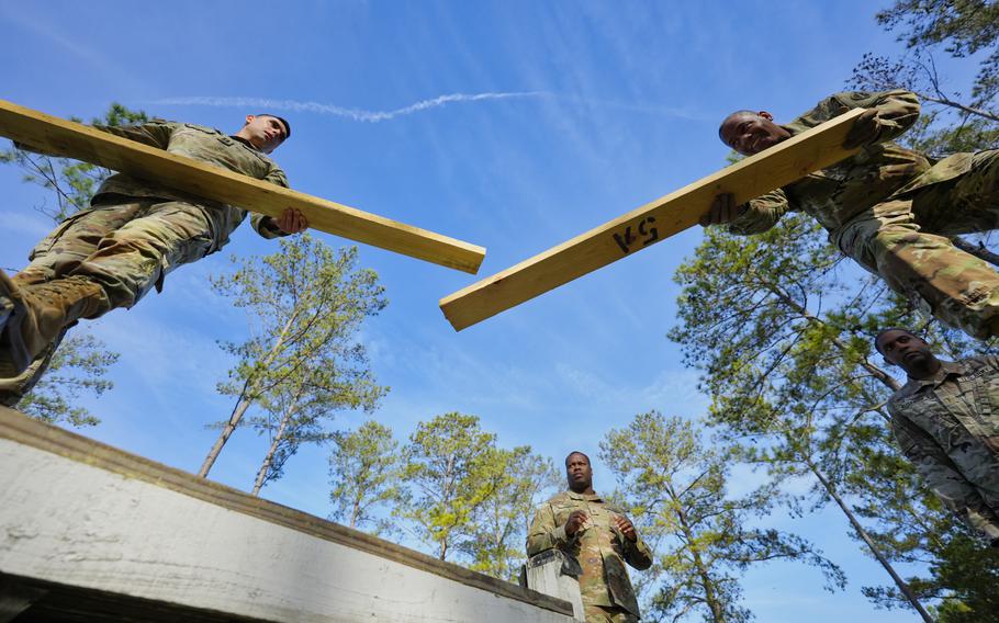 U.S. Army soldiers in camouflage uniforms participate in a team-building exercise outdoors, balancing wooden beams between platforms under a clear blue sky, surrounded by tall pine trees. 