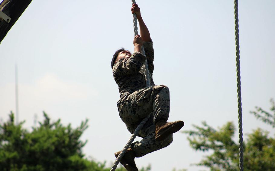 A U.S. service member in a camouflage uniform and tan boots climbs a thick rope during an outdoor obstacle course, surrounded by green trees under a bright sky.