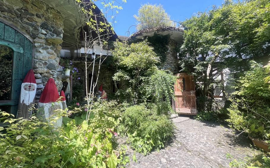 A stone path leads to a wooden door in a small plant-covered buildings, with wooden gnomes among plants on either side of the path.