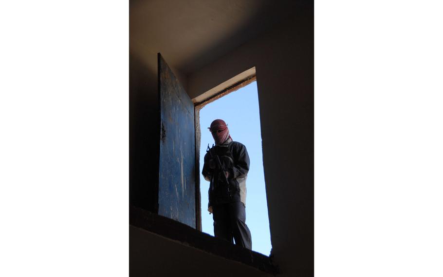 An armed and scarved man stands in a door frame, seen from below.