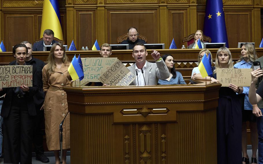 A man in a gray blazer sits at a large wooden podium inside the Ukrainian parliament building as supporters holding signs stand around him.