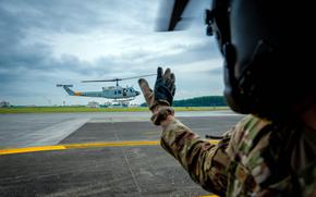 A flight crew member in camouflage uniform, gloves and flight helmet, seen from behind, waves to a helicopter as it takes off from an air base runway.