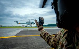 A flight crew member in camouflage uniform, gloves and flight helmet, seen from behind, waves to a helicopter as it takes off from an air base runway.