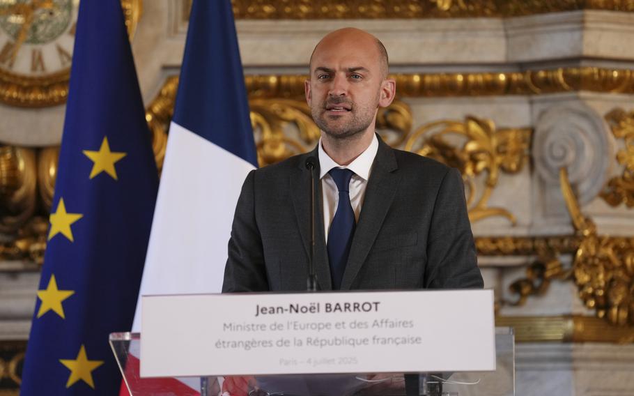 A man speaks at a lecturn in front of flags.