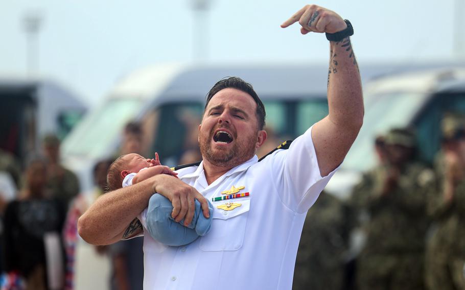 A sailor shouts happily, pointing at his baby, while celebrating his return from deployment.