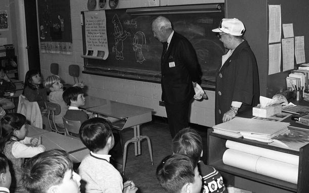 A black and white photo of two adults standing by a blackboard as young children sit at desks and watch the adults.