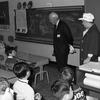 A black and white photo of two adults standing by a blackboard as young children sit at desks and watch the adults.