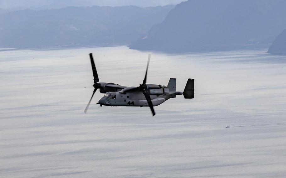 A military Osprey aircraft flies over open water with a mountainous coastline in the background.
