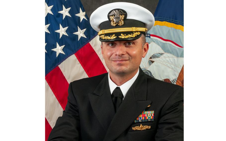 A naval officer in dress uniform and hat poses in front of a U.S. flag.