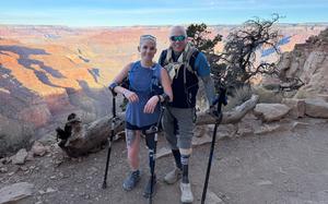 A man and a woman, each wearing hiking gear and holding crutches to support prosthetic legs, pose on a trail in front of a Grand Canyon vista.