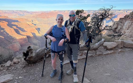 A man and a woman, each wearing hiking gear and holding crutches to support prosthetic legs, pose on a trail in front of a Grand Canyon vista.