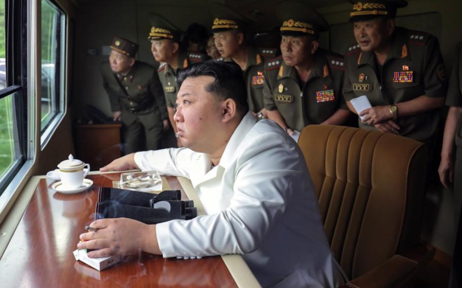 Kim Jong Un sits at a table and observes a weapons test through a window, with a line of military officials in green uniforms and hats standing behind him.