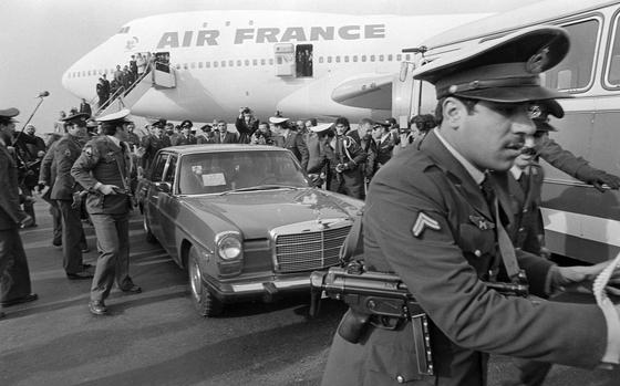 A security officer with a gun slung around his shoulder leans forward with his arms out to something off camera, as other security officers surround a car behind him.