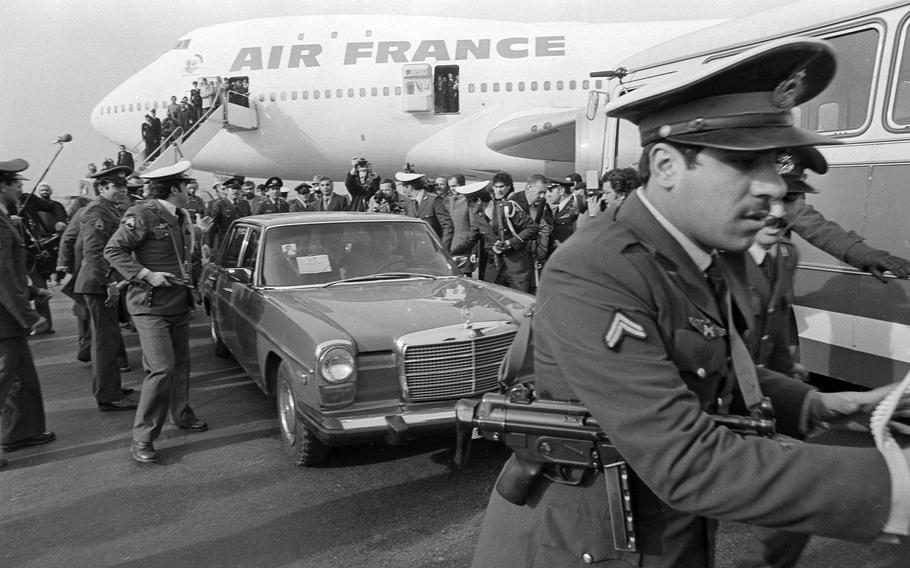 A security officer with a gun slung around his shoulder leans forward with his arms out to something off camera, as other security officers surround a car behind him.