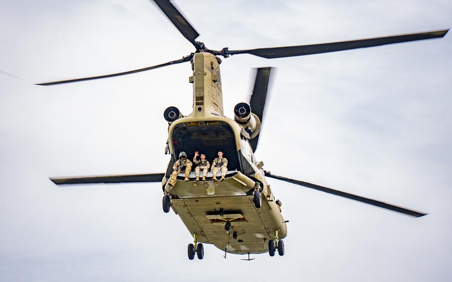 Three soldiers in a helicopter flying overhead, with one waving to people below.