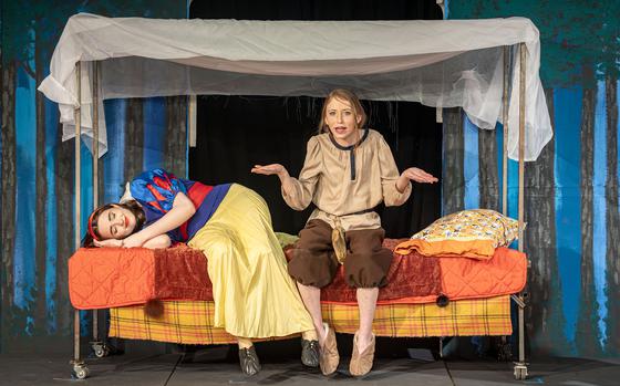 Snow White sleeps as a dwarf sits with her on a bed.