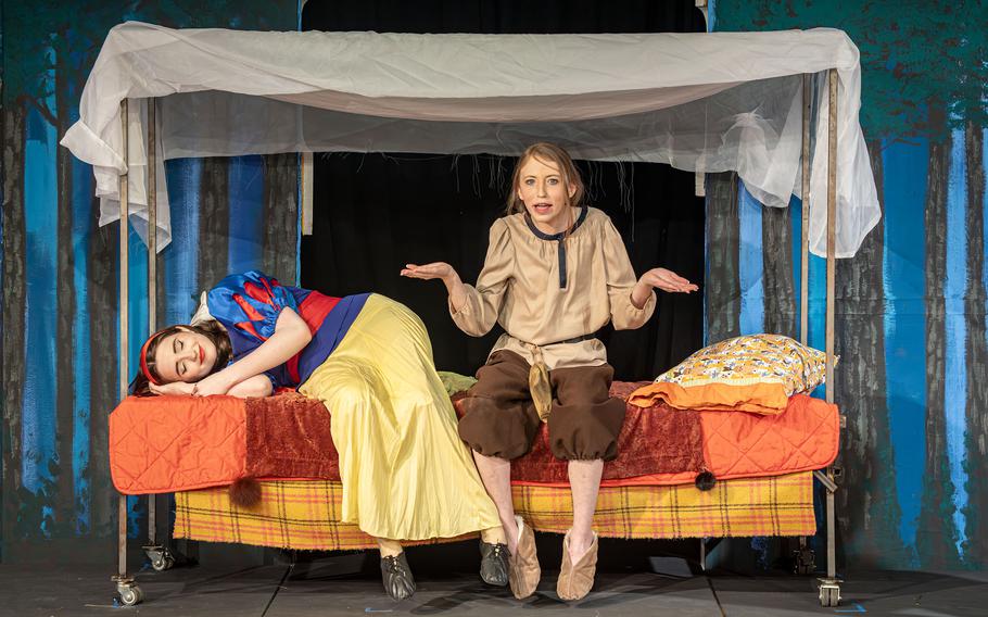 Snow White sleeps as a dwarf sits with her on a bed.