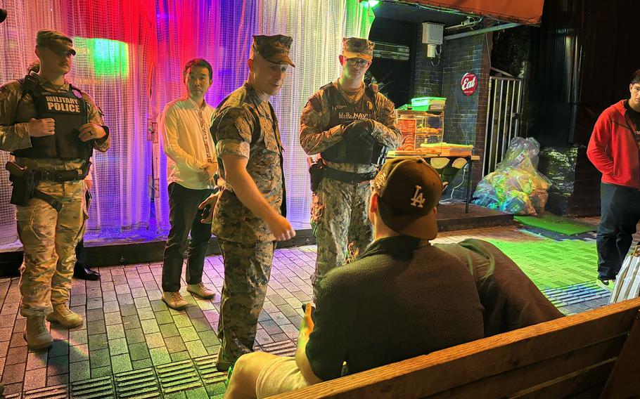 Military officials in camouflage uniforms speak to service members in civilian clothes seated on a street bench at night, near a colorful curtain backdrop.