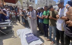 A line of Palestinian men with their arms linked stand and pray over a body covered by a white shroud laying on the dusty ground.