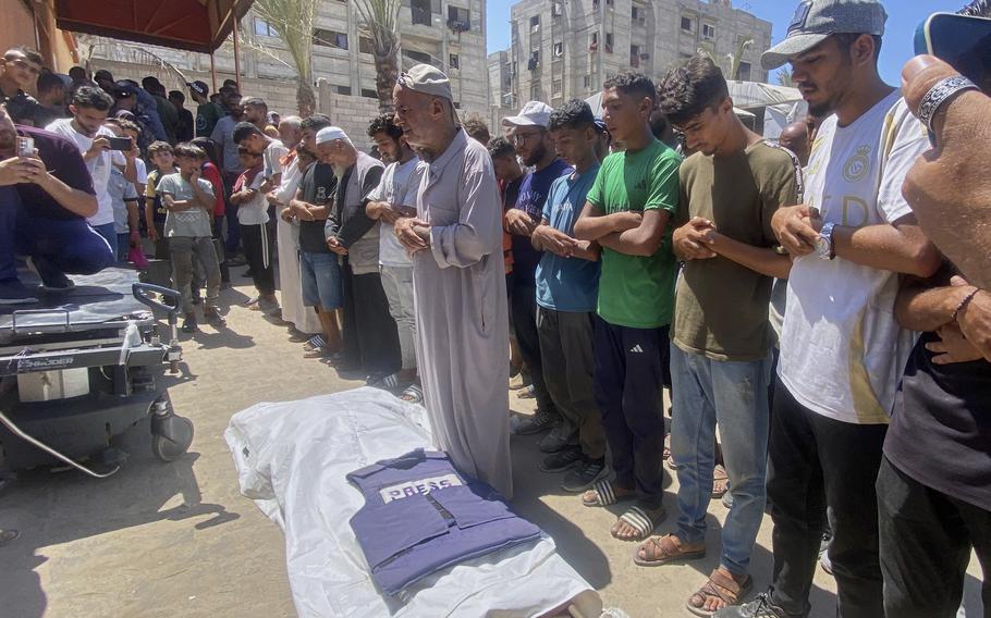 A line of Palestinian men with their arms linked stand and pray over a body covered by a white shroud laying on the dusty ground.