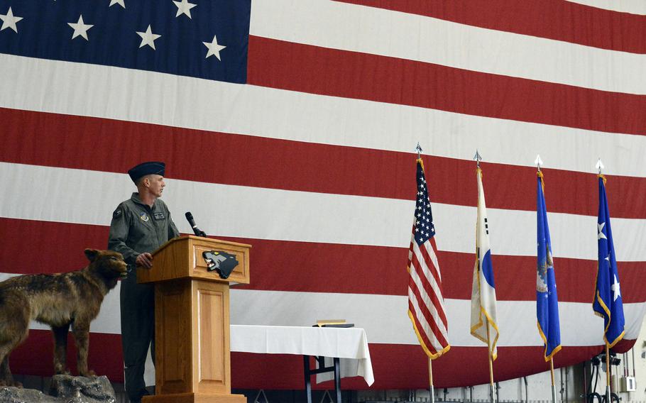 A military officer in green flight suit stands at a wooden podium speaking into a microphone. 