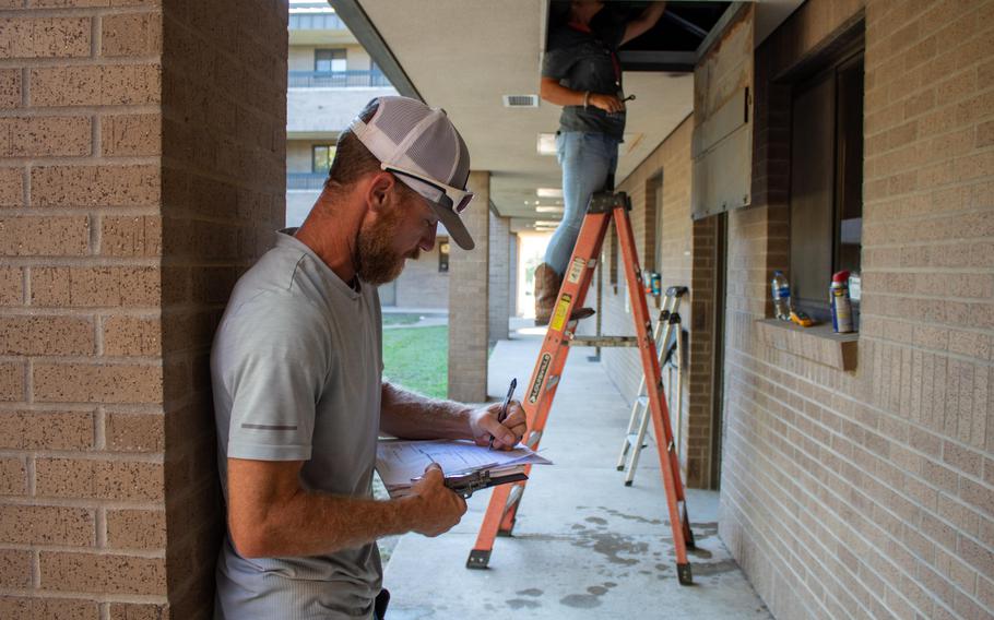 Two maintenance workers conduct repairs. One writes the paperwork as the other stands on a ladder and does the work.