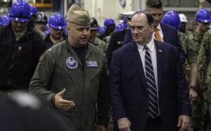 A man in a suit speaks with a military officer in a flight suit as they walk through a hangar bay in front of several employees wearing blue construction hard hats.