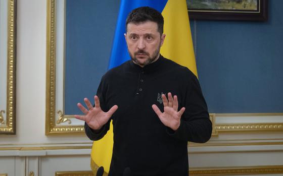 Volodymyr Zelenskyy raises his hands in gesture, palms open, while speaking in front of the Ukraine flag.