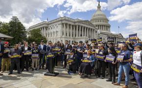 A senator stands at a podium and speaks while surrounded by rally-goers holding signs reading “Not For Sale,” with the U.S. Capitol building in the background.