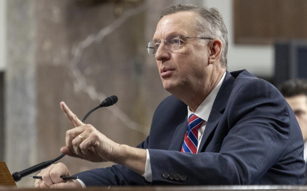 A man in a dark suit and glasses speaks into a microphone and points with his left hand while seated at a witness table in a congressional hearing room.