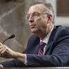 A man in a dark suit and glasses speaks into a microphone and points with his left hand while seated at a witness table in a congressional hearing room.