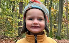 Caleb Anders, a toddler in a yellow jacket and dinosaur hat, poses for a photo in the woods.