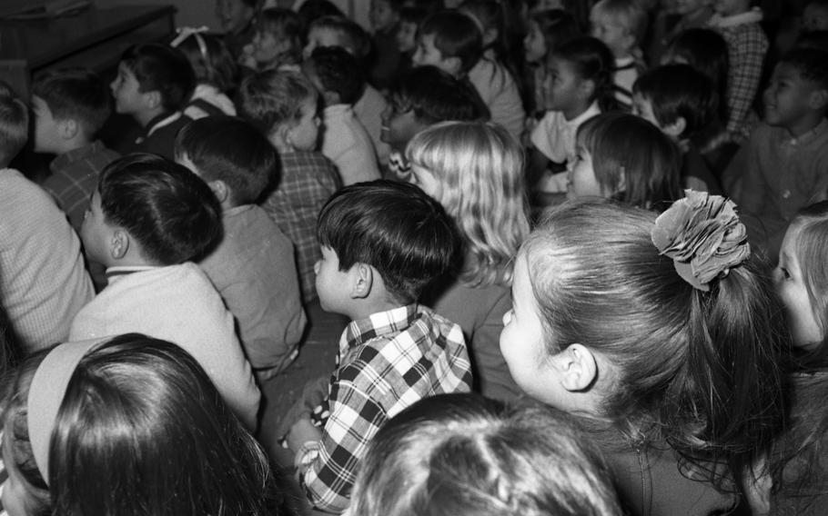 A black and white photo of school children seated and watching something.