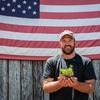 A man standing in front of an American flag holds an oregano seedling in his hands.